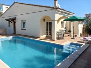 Un espace aménagé au bord de la piscine, comprenant une piscine, un patio couvert avec une table et des chaises, ainsi qu’un petit jardin avec un barbecue.