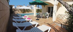 Un espace aménagé au bord de la piscine, avec des chaises longues, une table et des chaises, et un parasol vert.