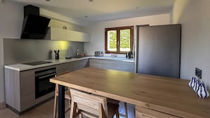 A modern kitchen with a wooden table, bar stools, a built-in oven, a refrigerator, and a window with a view of greenery.