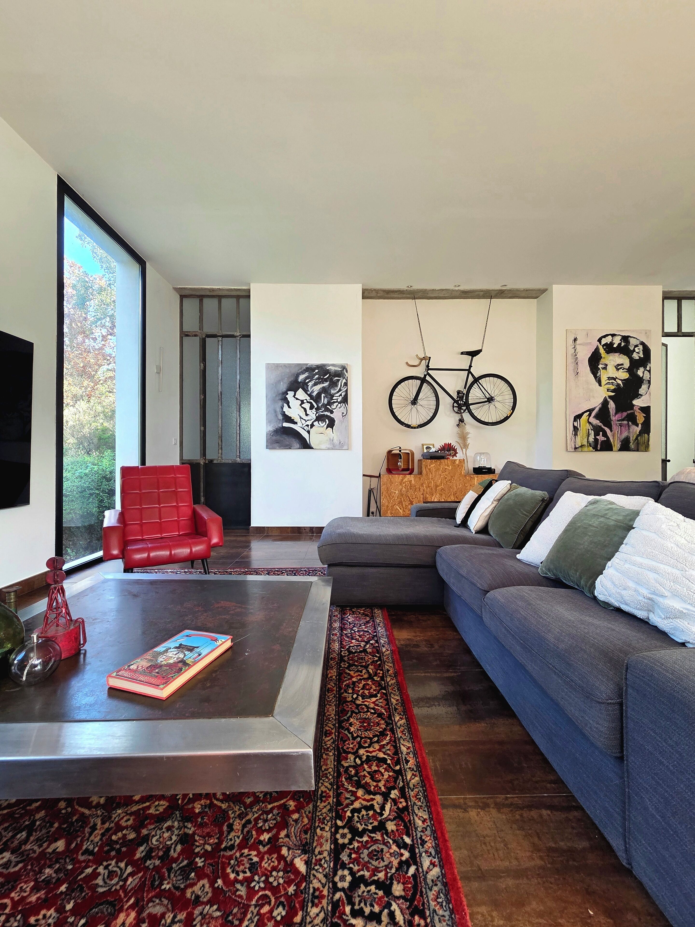 Un salon moderne avec un grand canapé d’angle gris, un fauteuil rouge, une table basse et une grande fenêtre.