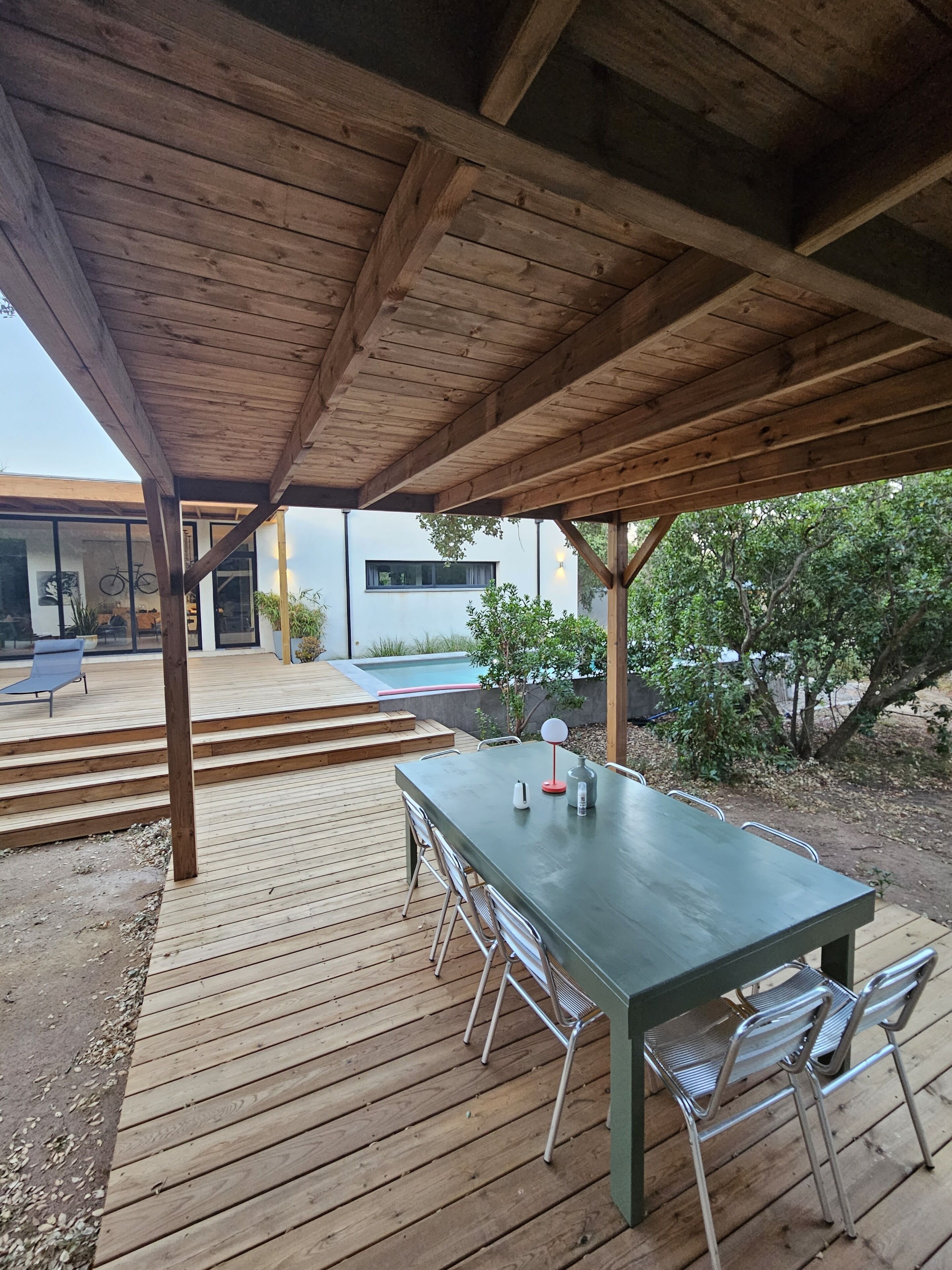 Une terrasse extérieure en bois, avec une table et des chaises, une piscine et un espace couvert offrant une vue sur un bâtiment moderne.
