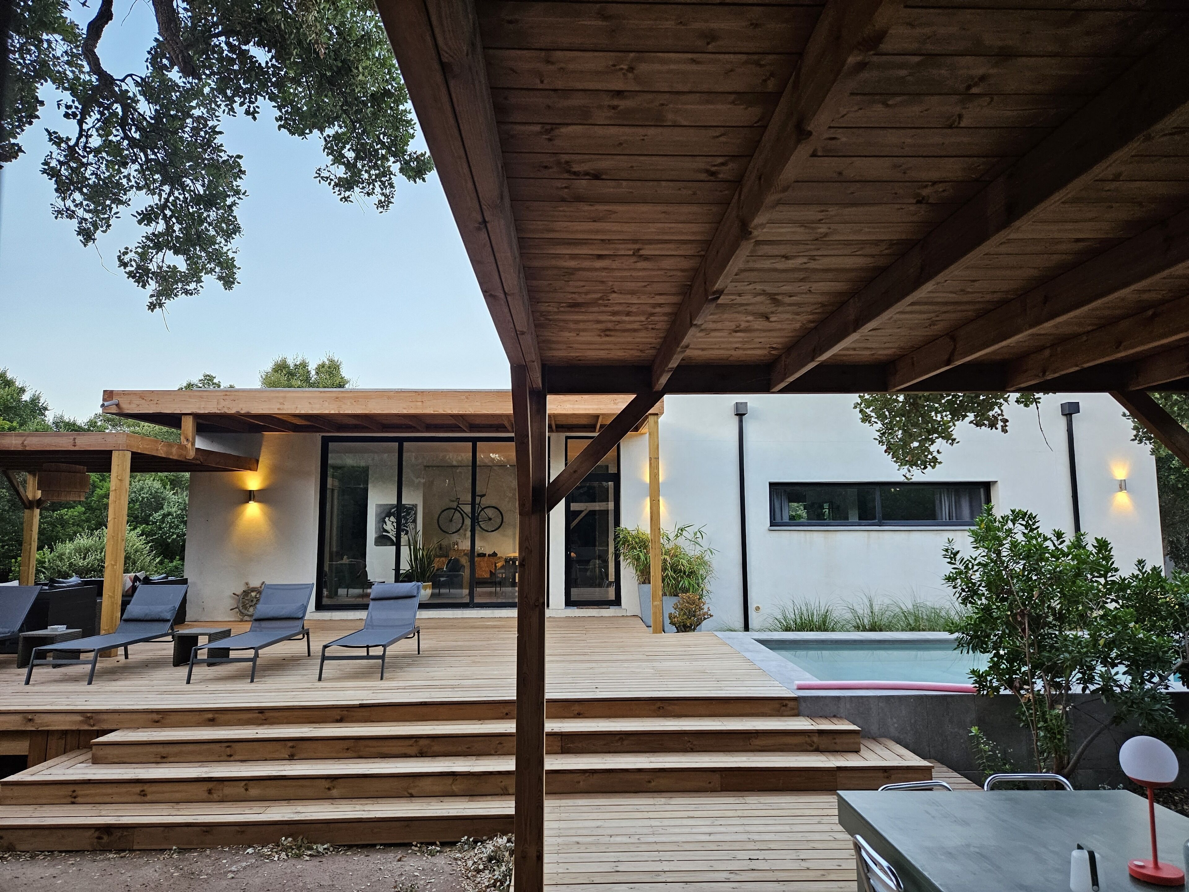 Een modern terras buiten met houten vloer, een zwembad en een overdekte zithoek met loungestoelen.