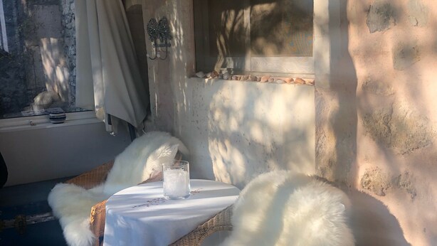 A room with a wicker chair and a table, a window with a view of the outside, and a wall with peeling paint. - Charming old village house, very quiet, wooded garden, sleeps 4. (Paradou)