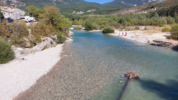 A river with clear water, surrounded by rocky terrain and greenery. There are people near the riverbank, and a vehicle parked on the left.