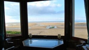 Une pièce dotée d’une grande fenêtre donnant sur une plage. On y trouve une table à plateau de verre, accompagnée de deux chaises, ainsi qu’une petite table sur laquelle sont posées deux bougies.