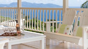 Eine Terrasse mit weißer Möbel, einer Weinflasche und Gläsern auf einem Tisch, mit Blick auf eine malerische Berg- und Meereslandschaft.