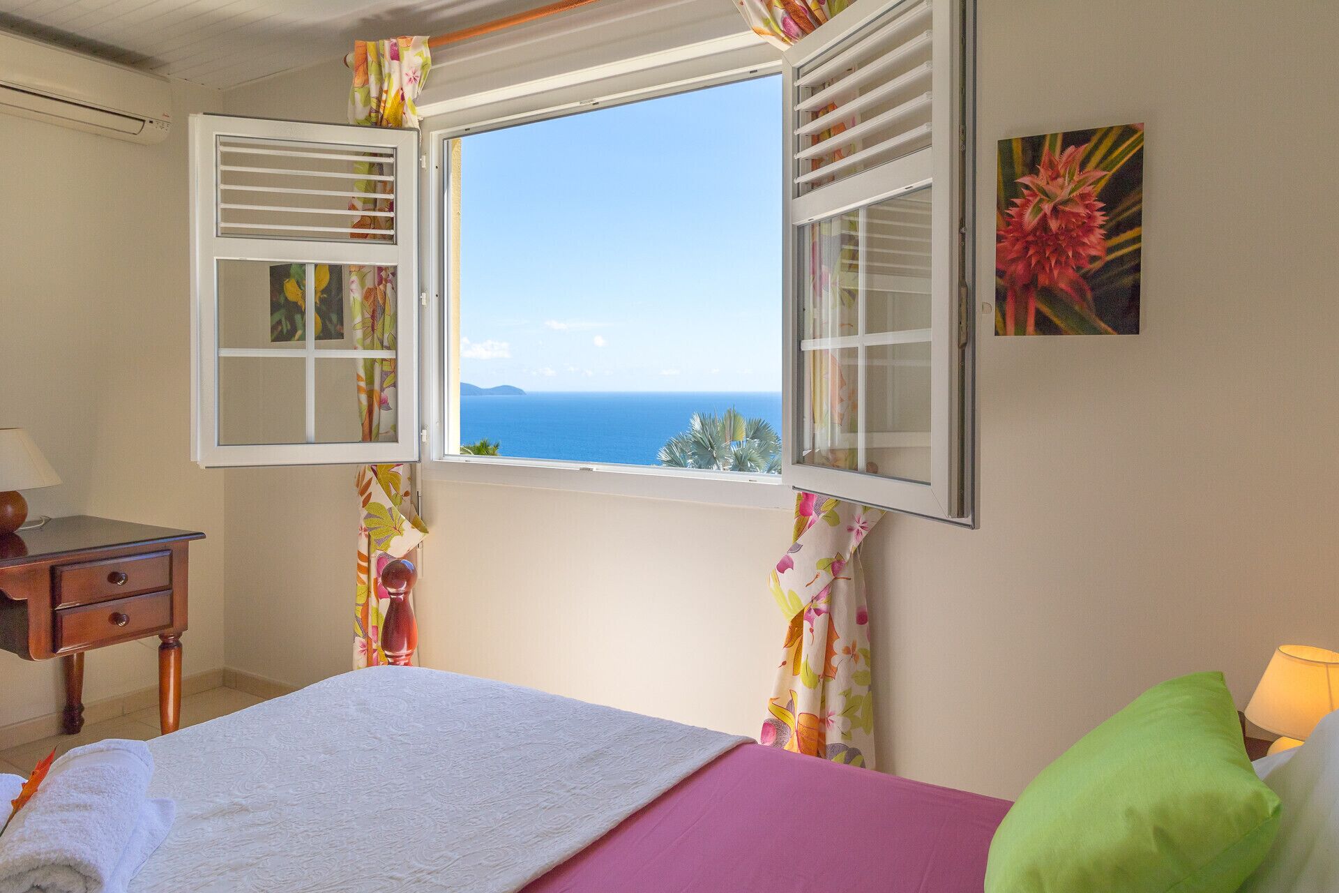 A bedroom with a bed, a nightstand, a lamp, and a window with a view of the sea.