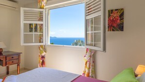 Ein Schlafzimmer mit einem Bett, einem Nachttisch, einer Lampe und einem Fenster mit Blick auf das Meer.