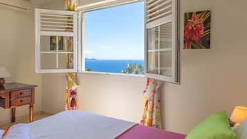 A bedroom with a bed, a nightstand, a lamp, and a window with a view of the sea.