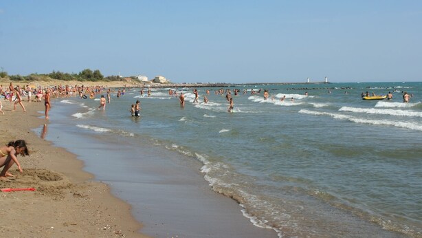 Une plage où l’on voit des gens se baigner et jouer dans l’eau, un rivage sablonneux et un phare au loin.