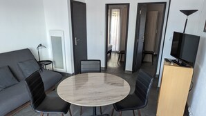 A modern living room with a grey sofa, a round wooden table, and black chairs. There is a TV on a wooden cabinet and a lamp on a side table. - Appt Leucate- La Franqui.  3 rooms, beach at 50 m, Terrace, free Wifi, (Leucate)