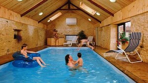 Una piscina interior con vigas de madera y paredes de piedra, rodeada de sillones de descanso y un pequeño jardín.