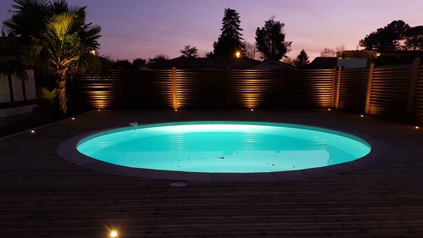 A circular swimming pool with a wooden deck surrounding it, illuminated by ambient lighting.