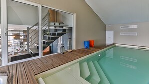 En moderne indendørs swimmingpool med en træterrasse, en glasdør der fører til en trappe, og udsigt over rummet indeni.
