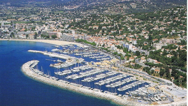 Une vue aérienne d’une ville côtière, avec un port de plaisance rempli de bateaux, entourée d’immeubles résidentiels et dominée, à l’arrière-plan, par un paysage vallonné.