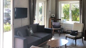Un soggiorno moderno con un divano grigio, un tavolino in legno e una poltrona bianca. C'è una TV a schermo piatto montata a parete, e la stanza ha ampie finestre con tende.