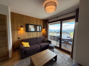 A modern living room with a wooden wall, a dark sofa, a wooden coffee table, and a view of mountains through large sliding doors.