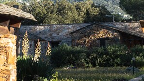 Un bâtiment en pierre doté d’un toit en pente, entouré de verdure et avec une montagne en arrière-plan.