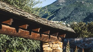 Une construction en pierre surmontée d’un toit en bois, entourée de verdure et avec une montagne en arrière-plan.