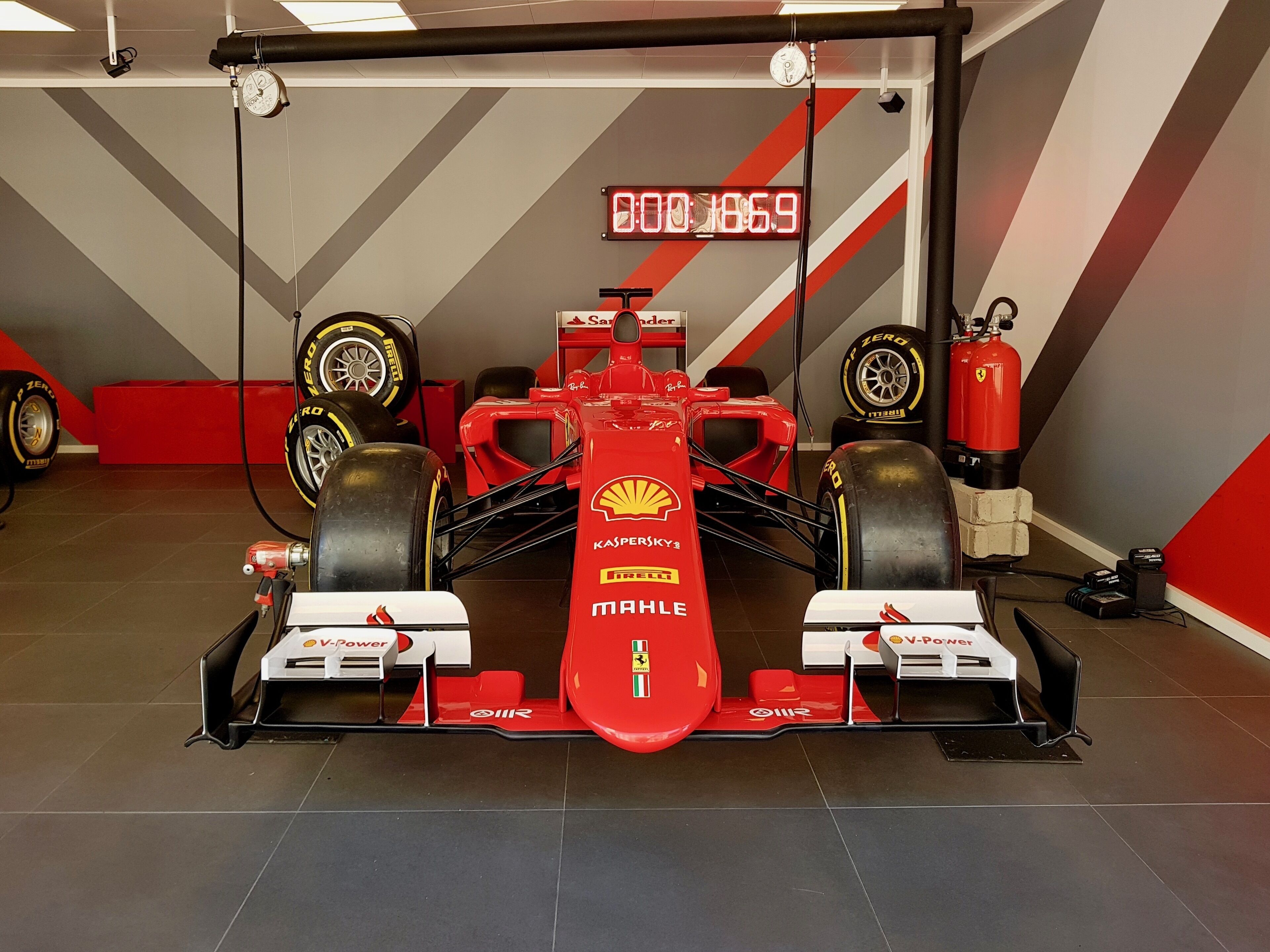 A red Formula 1 car with sponsor logos, tires, and a digital clock in a garage.