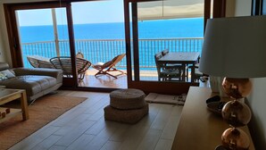 Un salon comprenant un canapé, une table basse, un pouf en osier et une vue sur l’océan, visible à travers une porte-fenêtre coulissante.