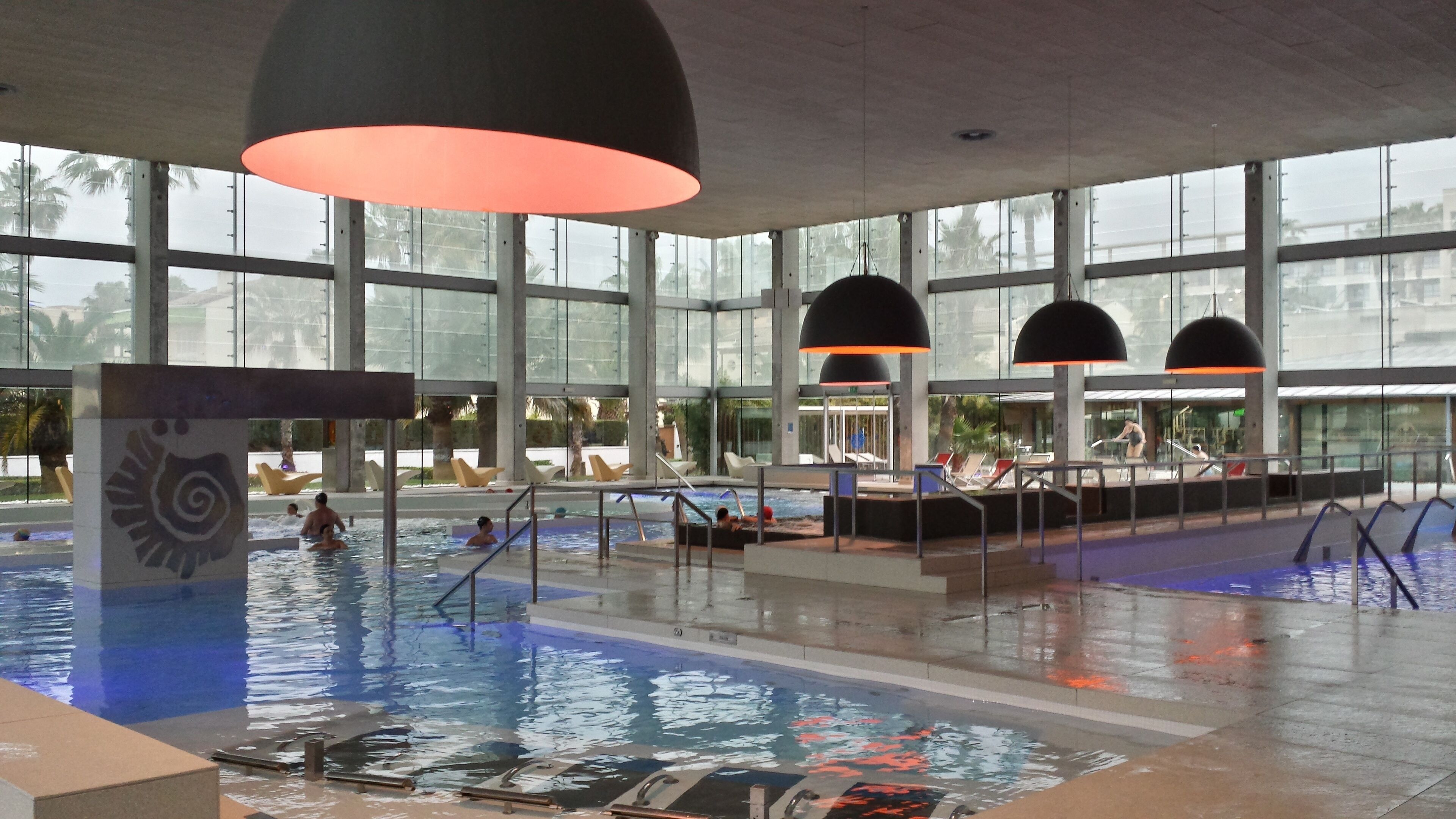Una piscina interior con paredes de vidrio transparente, varias tumbonas y grandes lámparas colgantes.