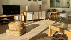 Un salon moderne avec un téléviseur à écran plat, un meuble en bois, un pouf en fibres naturelles, une table basse en verre avec des magazines, et un coin repas avec des chaises et une table.