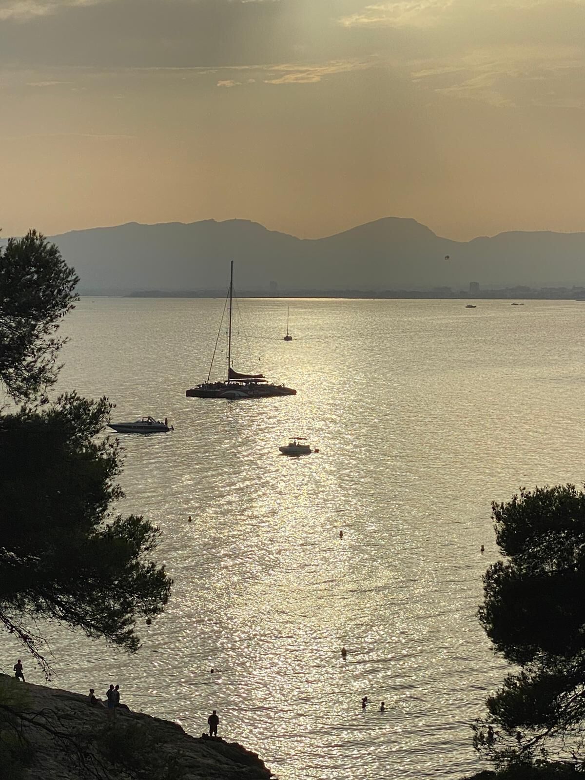 A calm body of water with boats and a sailboat, surrounded by trees and mountains in the background.