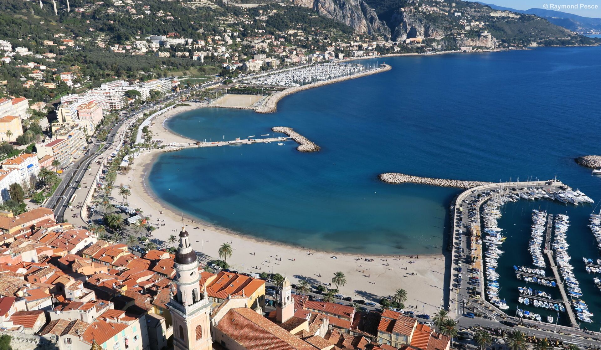 An aerial view of a coastal town with a marina, beach, and residential buildings.