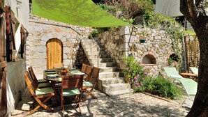 Ein Steinhaus mit einer Holztür und einer Treppe, die zu einer oberen Ebene führt. Ein Tisch mit Stühlen und ein grüner Sonnenschirm spenden Schatten.
