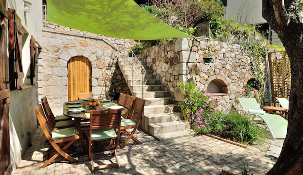 A stone building with a wooden door and a staircase leading to an upper level. There is a table with chairs and a green umbrella providing shade.