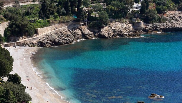 Ein Küstenstrand mit klarem türkisblauem Wasser, Felsformationen und einem gepflasterten Gehweg.