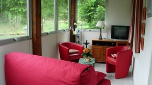 A room with a red sofa, two red chairs, a small table with a lamp, and a television on a wooden cabinet. - Chalet overlooking a pond in a green setting. (Augignac)