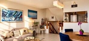 A modern living room with a sofa, coffee table, and artwork. A staircase leads to a kitchen area with a refrigerator and a dining table. - Stunning triplex with views of the ocean and the marina from the balcony, close to downtown (Deauville)