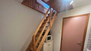 Uma escada de madeira leva a um mezanino, um armário branco com micro-ondas e uma porta rosa em um quarto com piso ladrilhado.
