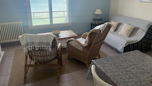 Quarto com vista para o mar, uma cadeira de vime, um sofá, uma mesa pequena e um abajur.