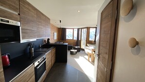 A modern kitchen with wooden cabinetry, a black countertop, and a built-in oven. There is a dining area with a table and chairs, and a large window providing natural light. - CHARMING APARTMENT 4 ROOMS 9 PEOPLE AVORIAZ (Morzine)