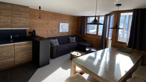 A modern kitchen with wooden cabinets, a black countertop, and a built-in oven. There is a dark-colored sofa with a coffee table and a glass dining table with white furry stools. - CHARMING APARTMENT 4 ROOMS 9 PEOPLE AVORIAZ (Morzine)
