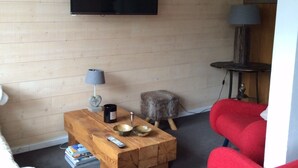 Una sala de estar con piso de madera, televisor montado, un sillón rojo, una mesa de centro con libros y un control remoto, una lámpara y un adorno de asta de ciervo en la pared.