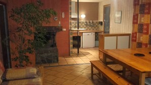 Un coin repas chaleureux avec une table et des bancs en bois, une cheminée en pierre et une cuisine en arrière-plan.