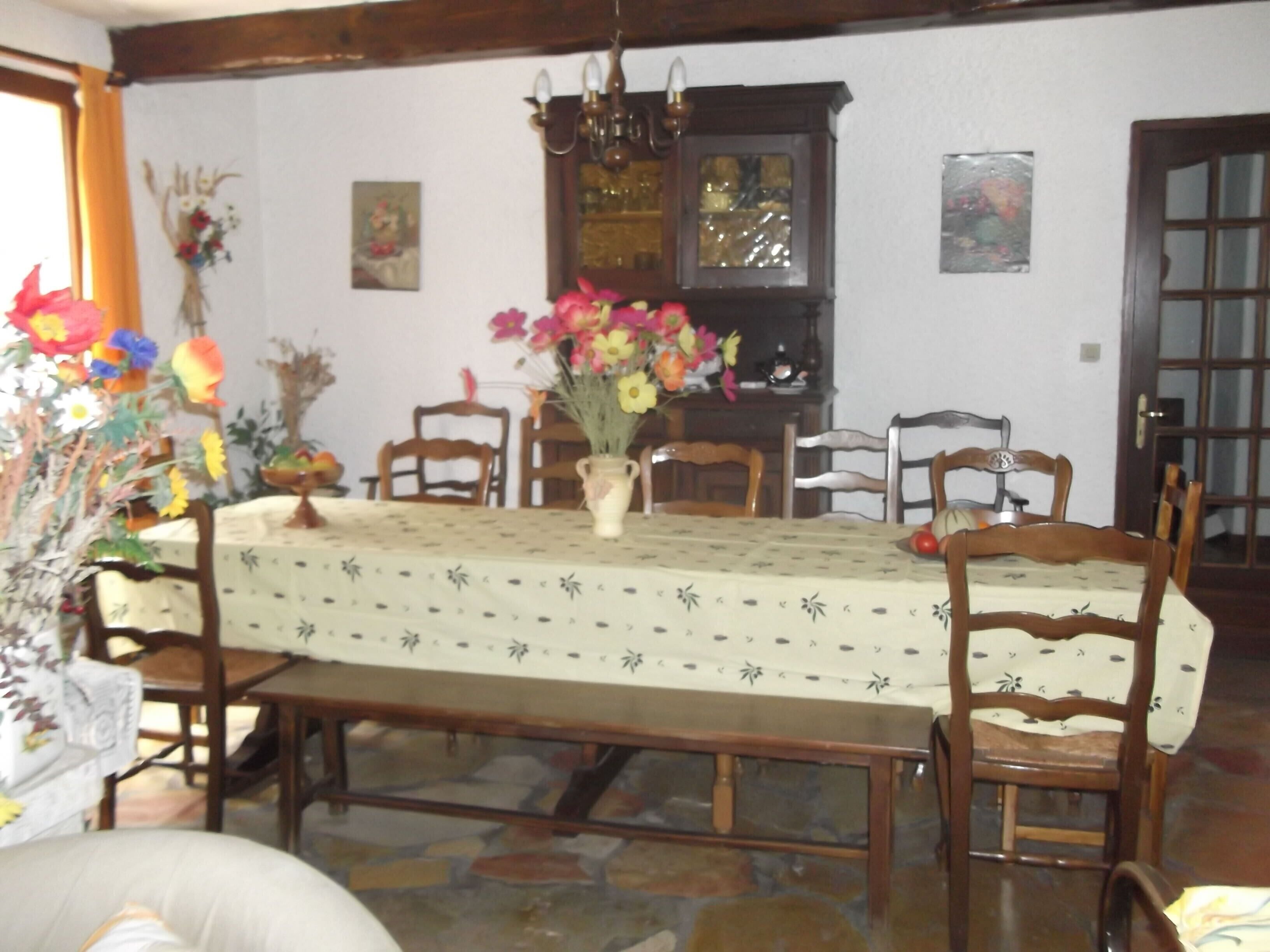A dining room with a long table, chairs, a vase of flowers, and a chandelier.