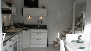 Cocina moderna con armarios blancos, encimera negra y horno empotrado. Hay un comedor con mesa y sillas blancas, y una estantería con libros y objetos decorativos.