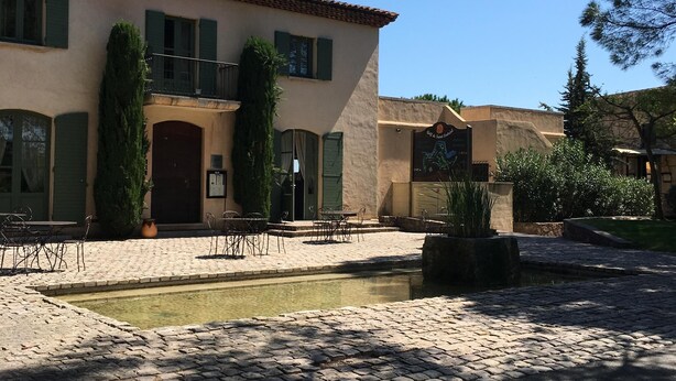 Un patio con piscina, mobiliario exterior y un edificio de techo a ladrillo y contraventanas verdes.
