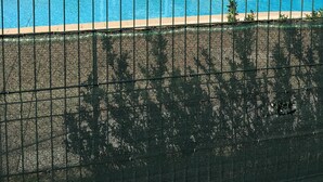 Una piscina con una valla en primer plano, un pérgola y árboles al fondo.