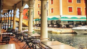 Un café al aire libre con mesas y sillas, un canal y edificios con balcones y contraventanas.