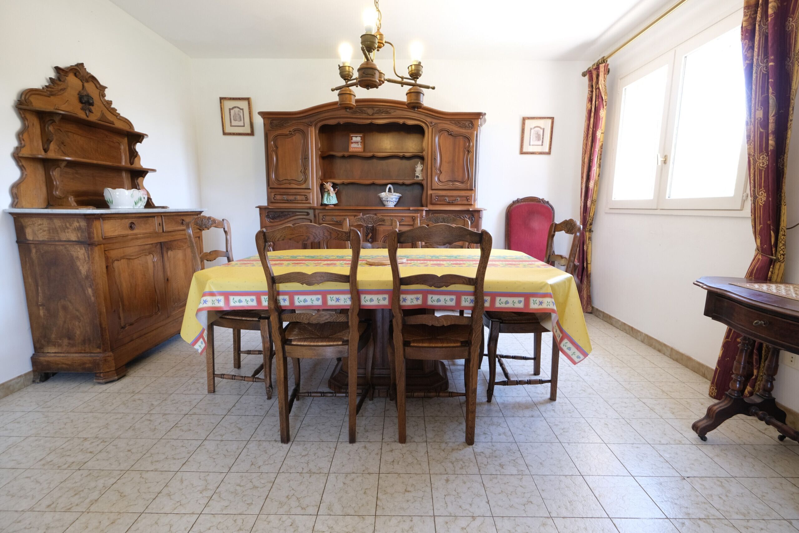 A dining room with a wooden table and chairs, a vintage wooden cabinet, and a window with curtains.