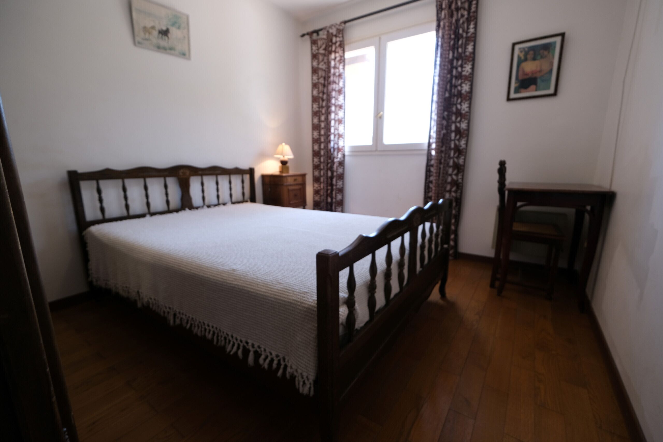 A bedroom with a wooden bed, a nightstand with a lamp, a window with curtains, and a small table.