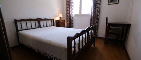 Un dormitorio con cama de madera, mesita de noche con lámpara, ventana con cortinas y una pequeña mesa.