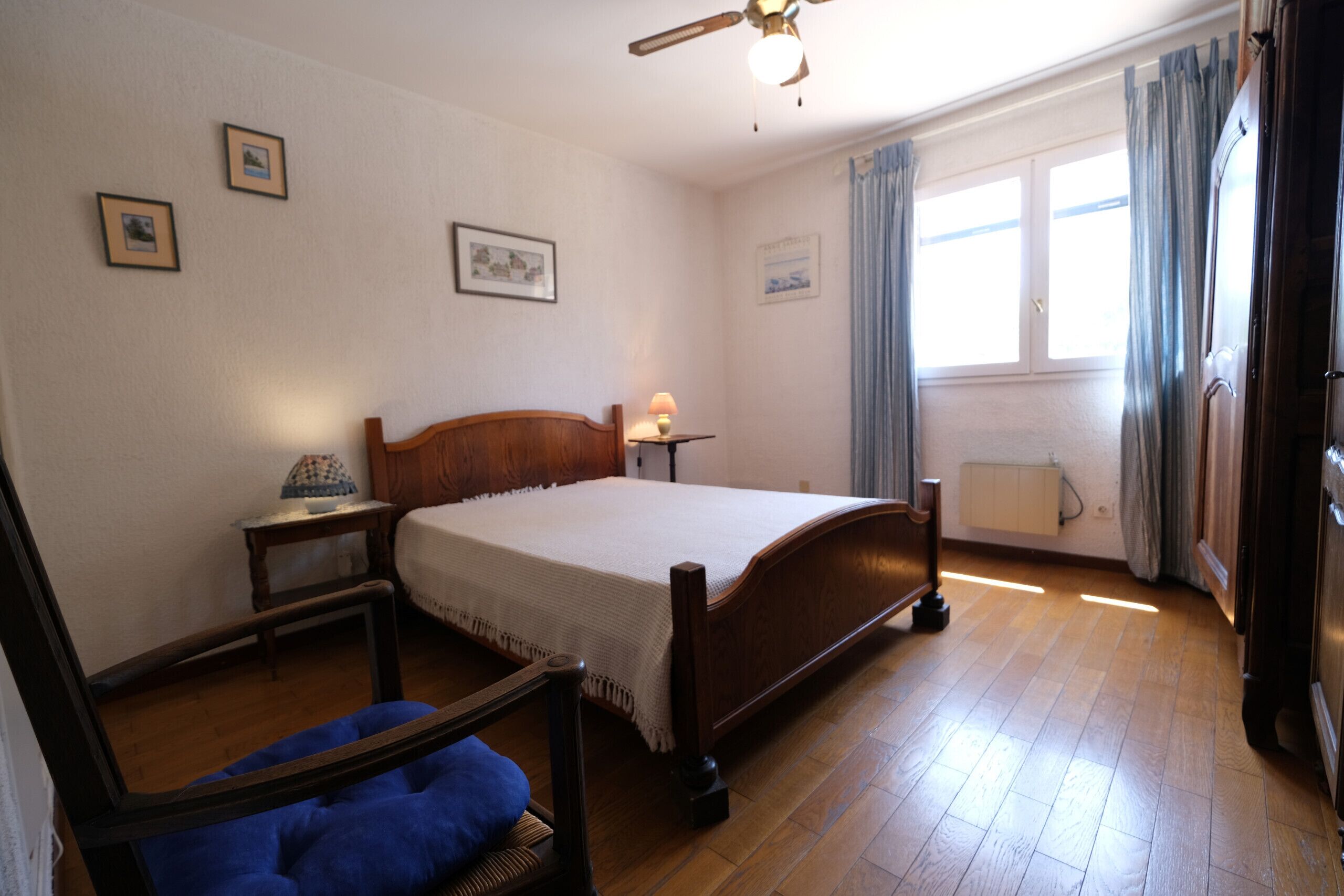 A bedroom with a wooden bed, a bedside table with a lamp, a chair, and a window with curtains.