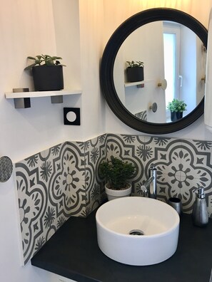 A bathroom with a white sink, black countertop, patterned tile backsplash, a round mirror, and two potted plants on floating shelves. - Charming Town House near the beaches of Normandy and Pegasus Bridge (Bénouville)
